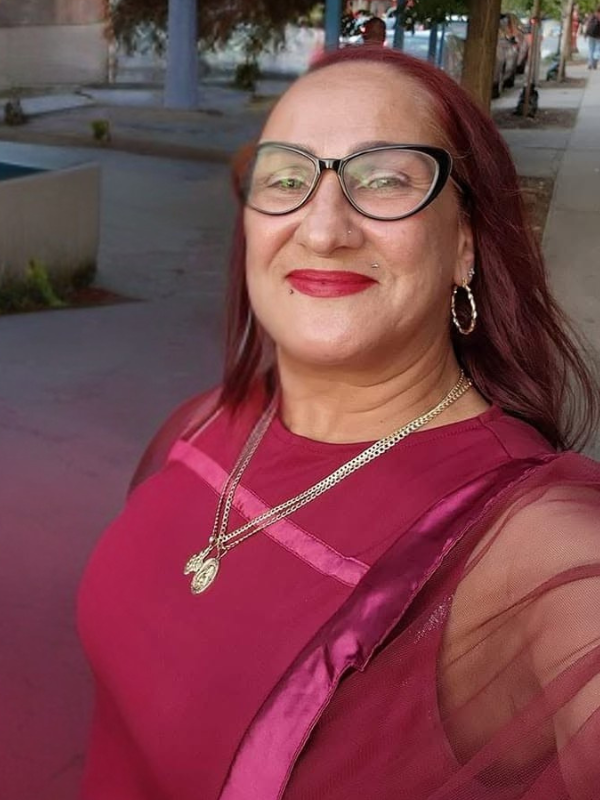 A woman with red hair, glasses, and earrings, smiling on a sidewalk with trees and parked cars in the background, wearing a dark pink top and gold jewelry.