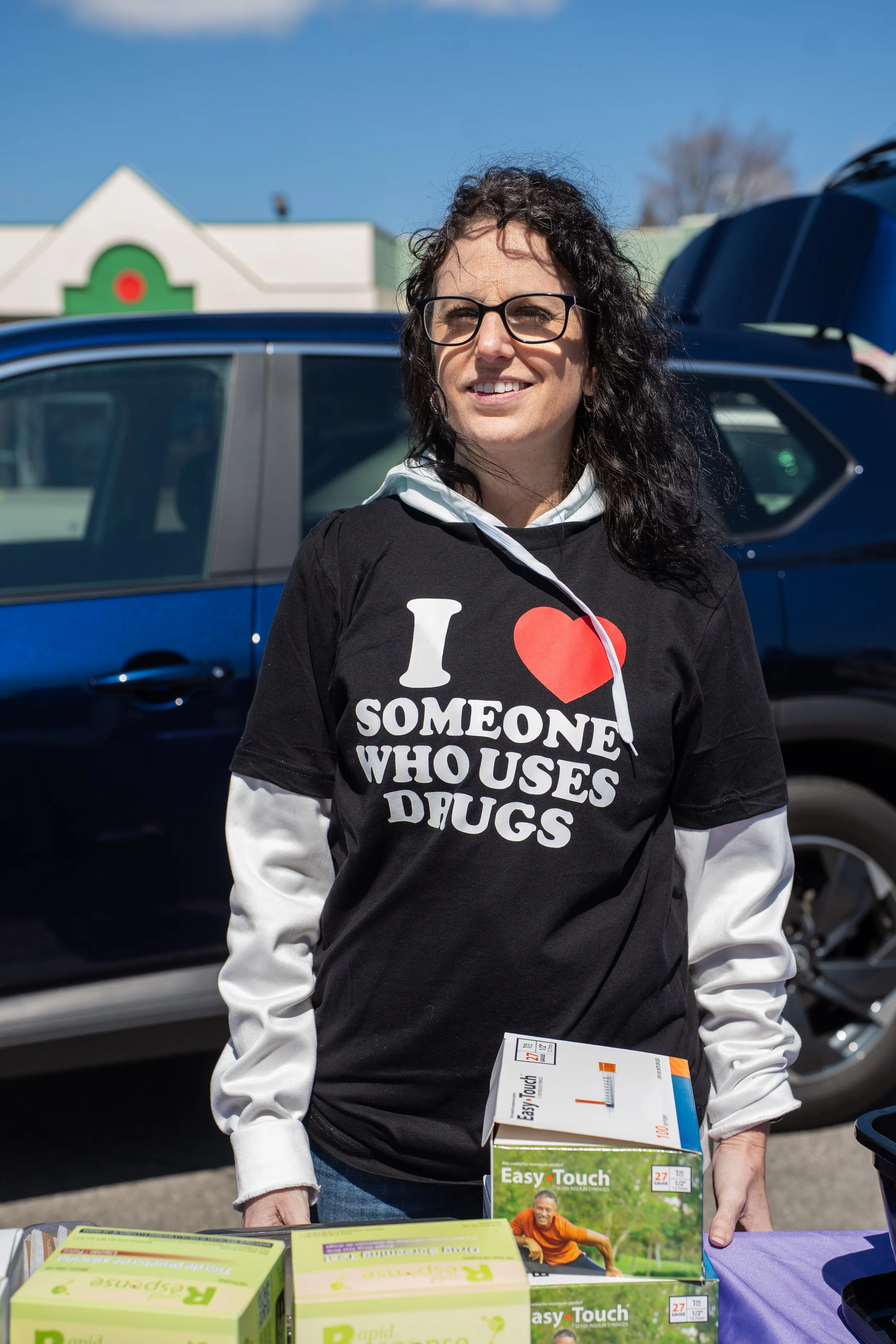 Woman with curly dark hair and glasses, wearing a black shirt with white and red text that says 'I ❤️ SOMEONE WHO USES DRUGS', standing outdoors at a table with health-related products.