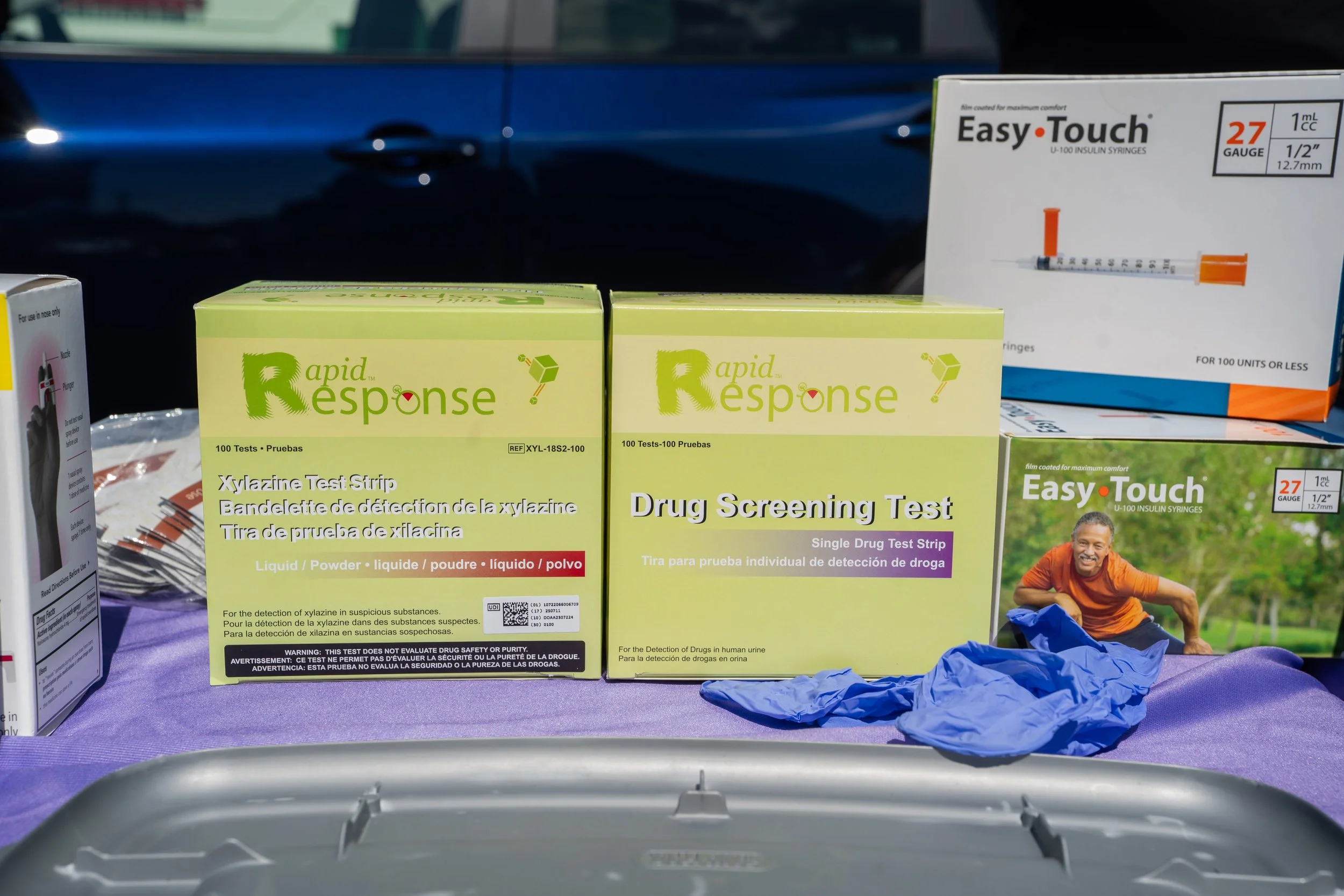Various drug testing kits and supplies displayed on a table, including boxes labeled 'Rapid Response Drug Screening Test' and 'Easy Touch' insulin syringes, with a person in an orange shirt and blue gloves in the background.