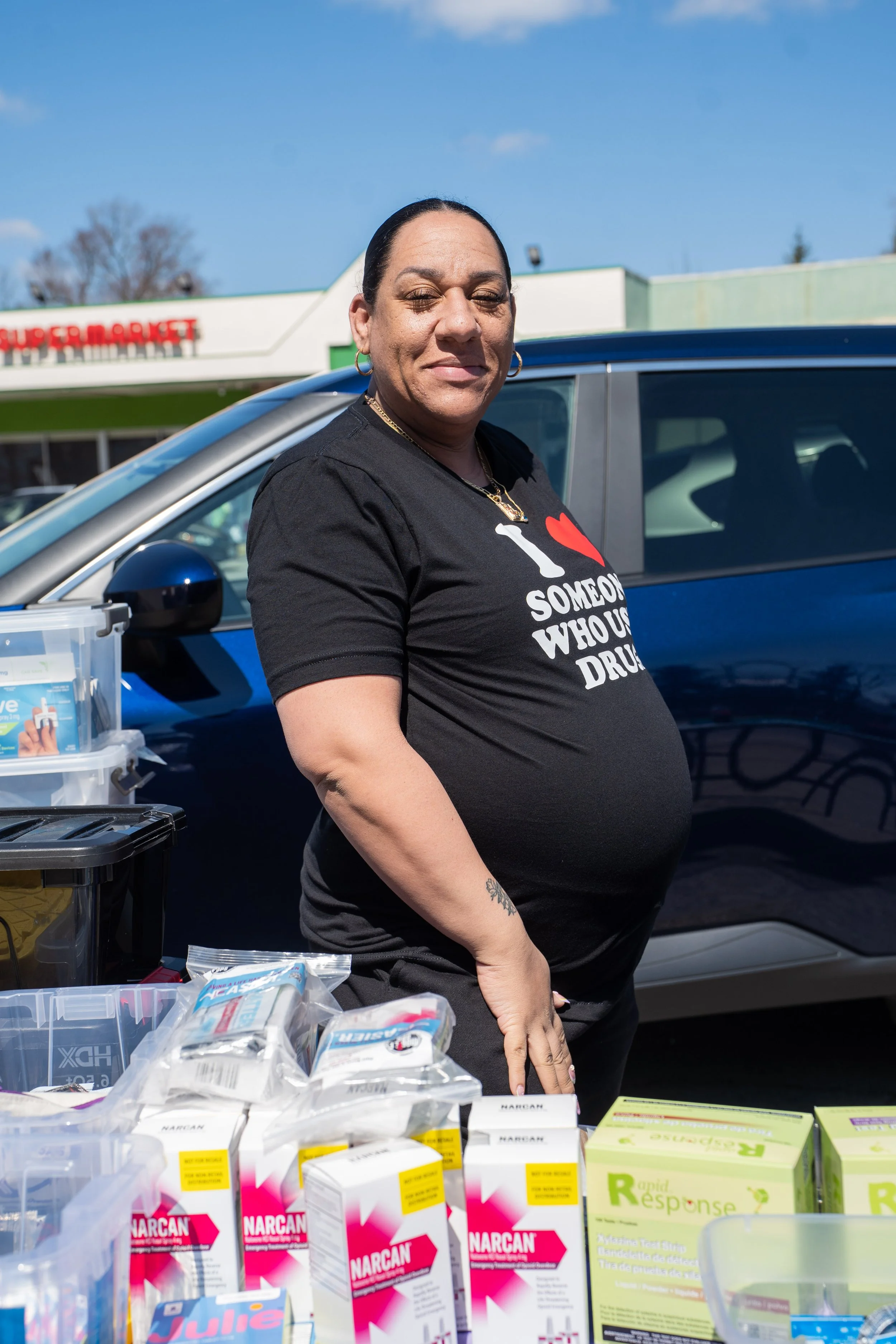 A woman standing outdoors near a vehicle, wearing a black T-shirt with a red heart and white text that reads 'I love someone who uses drugs.' She has dark hair pulled back, gold hoop earrings, and gold necklaces. In front of her are various medical supplies and drug testing kits on a table. In the background, there is a supermarket with a sign and a clear blue sky.