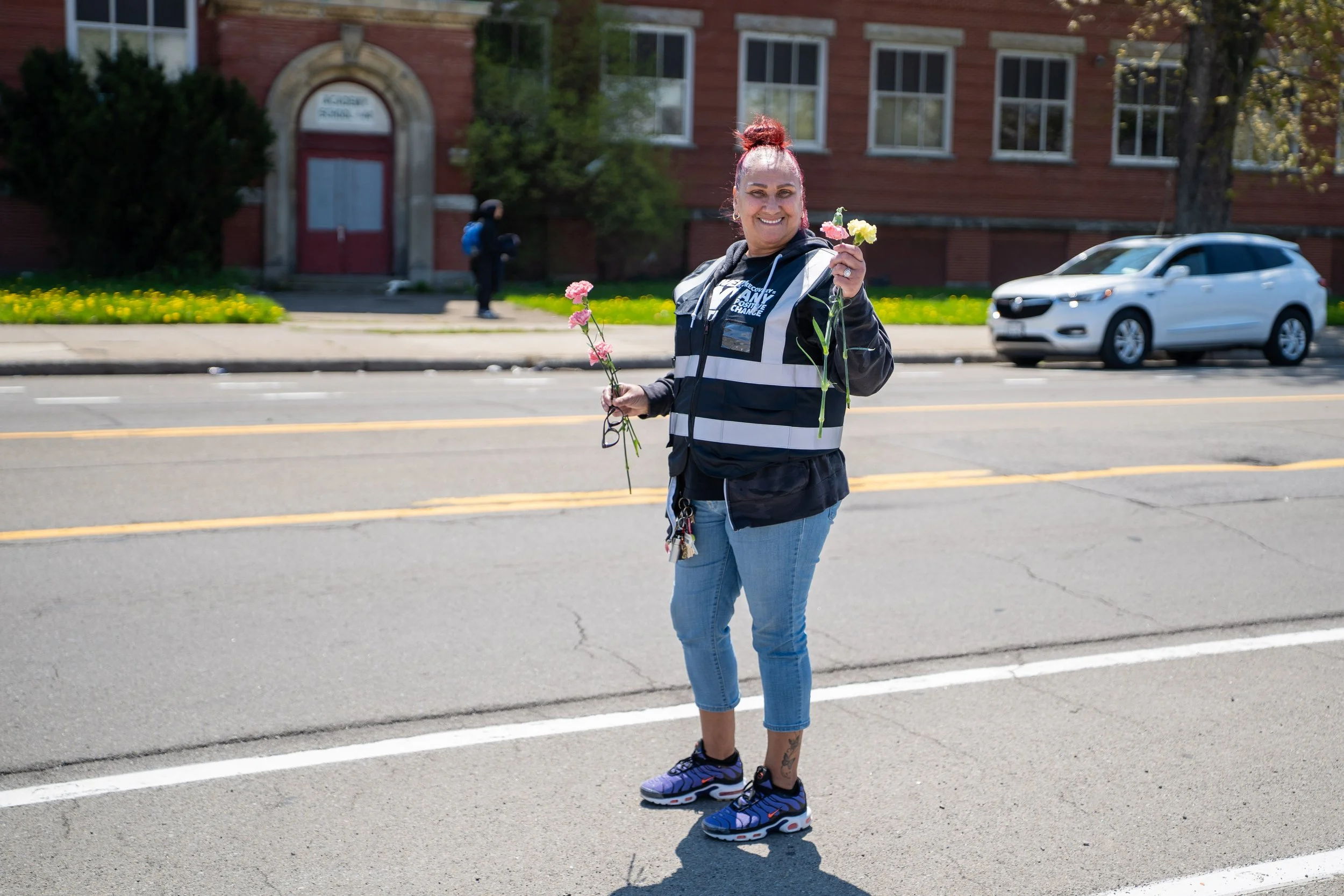 A woman with red hair in a bun, smiling, standing in the street holding pink and yellow carnations, wearing a black and white hoodie, jeans, and sneakers.