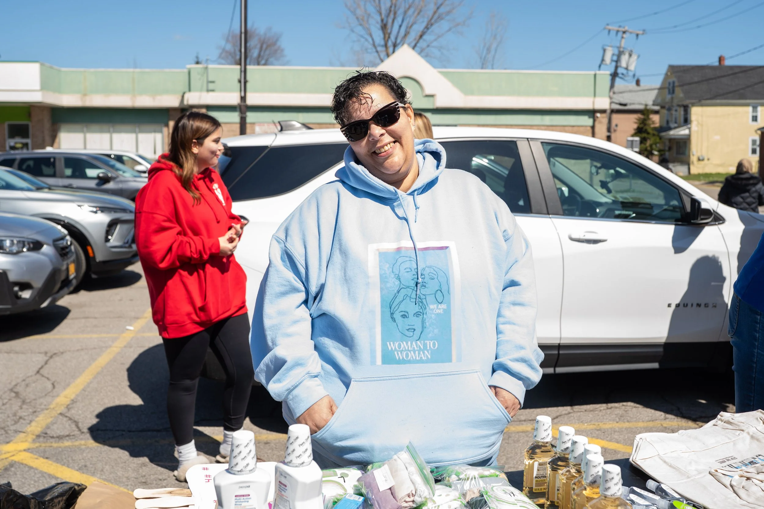 Woman with short dark hair and sunglasses smiling at camera, wearing a light blue hoodie. She is standing outdoors next to a table with various supplies, and there are others in the background including a woman in a red hoodie and several parked cars.