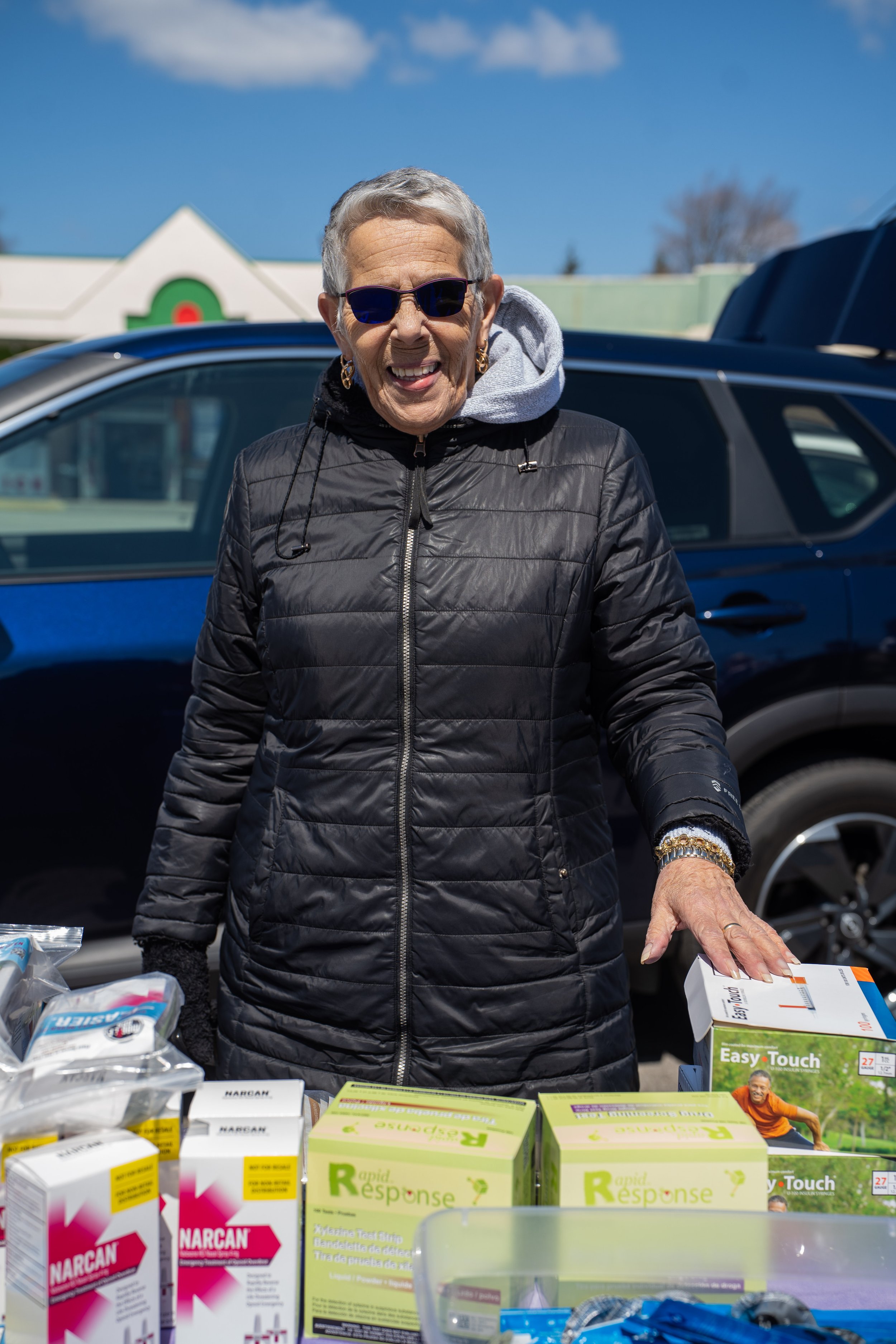 Smiling elderly woman with sunglasses and earrings handing over a box at an outdoor table with various health products, including Narcan and rapid response test kits, in front of a blue vehicle under a partly cloudy sky.