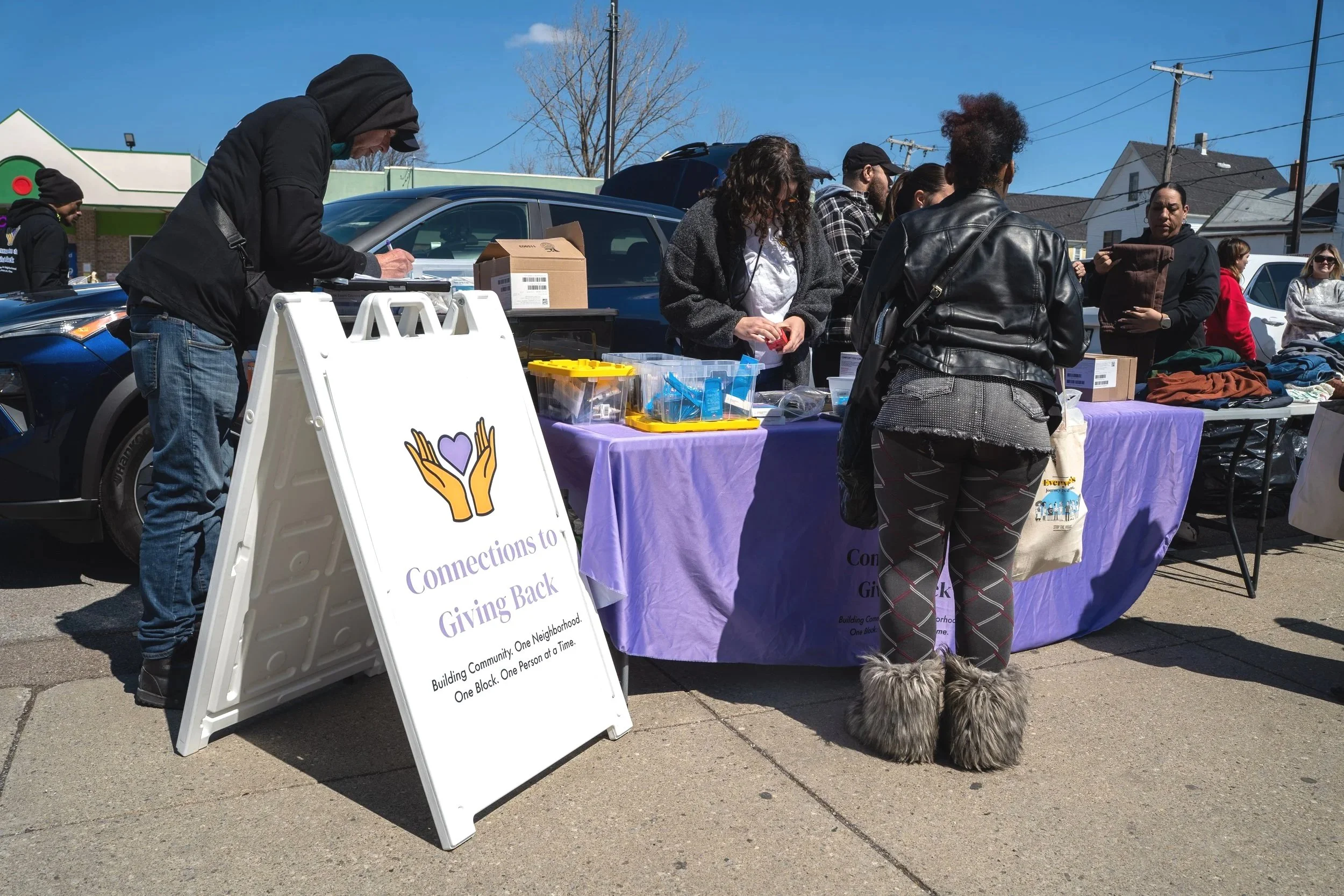 People standing behind a table at an outdoor event, with a sign that says "Connections to Giving Back" and supplies on the table, in a parking lot under a sunny sky.