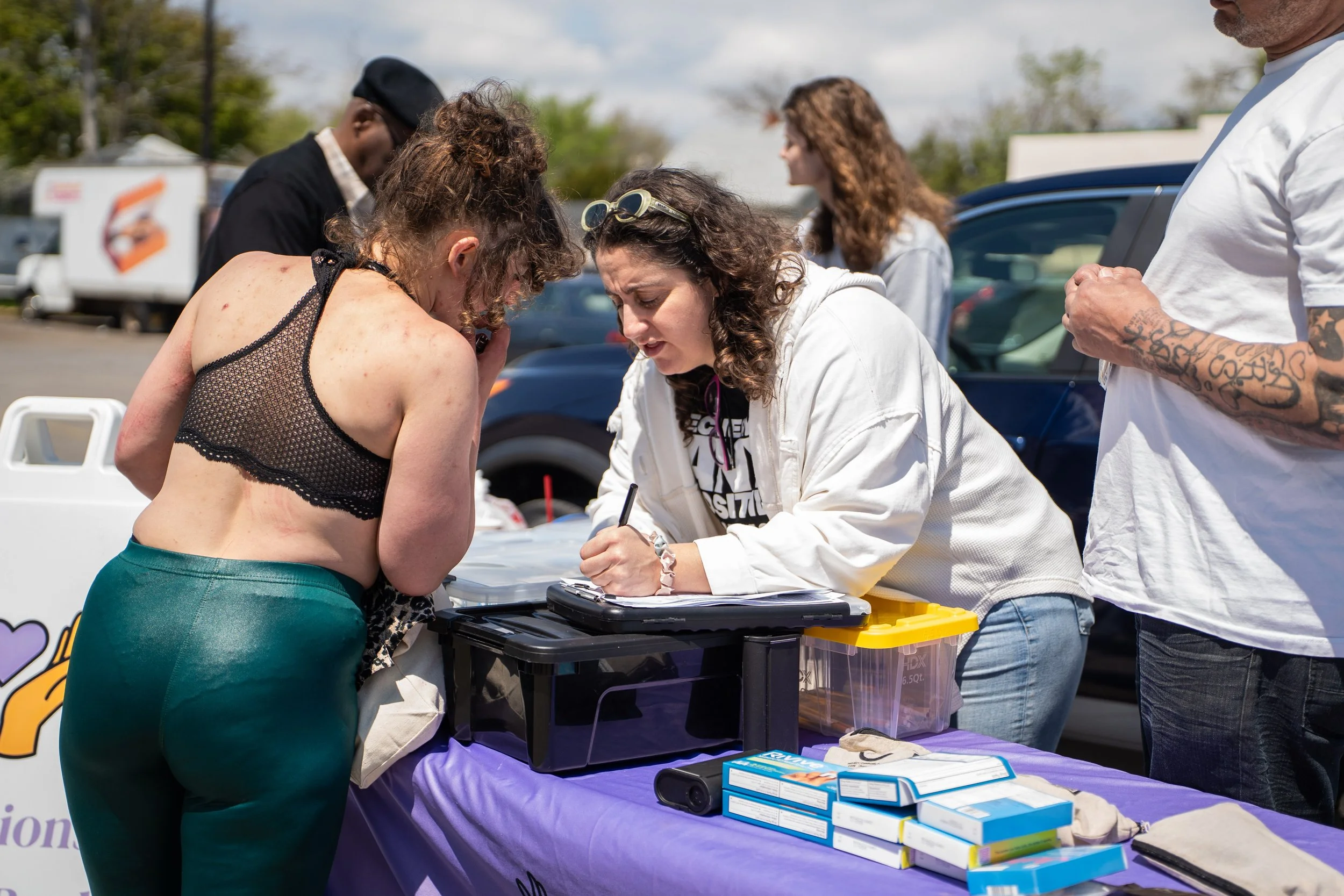 A woman in a patterned tank top and teal pants leaning over a table at an outdoor event, signing a document or waiver. Several people are standing around, and a woman with sunglasses and curly hair is assisting her. The table has medical supplies and paperwork, with a purple cloth covering it. There are cars parked in the background and a food truck.