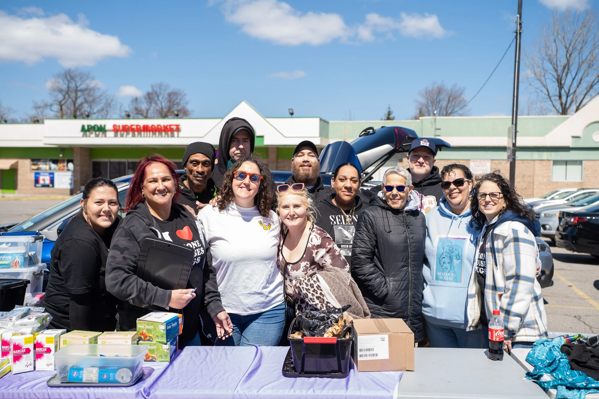 A diverse group of people standing outdoors at a community event in front of a supermarket, behind a table filled with various supplies and donations, under a partly cloudy sky.