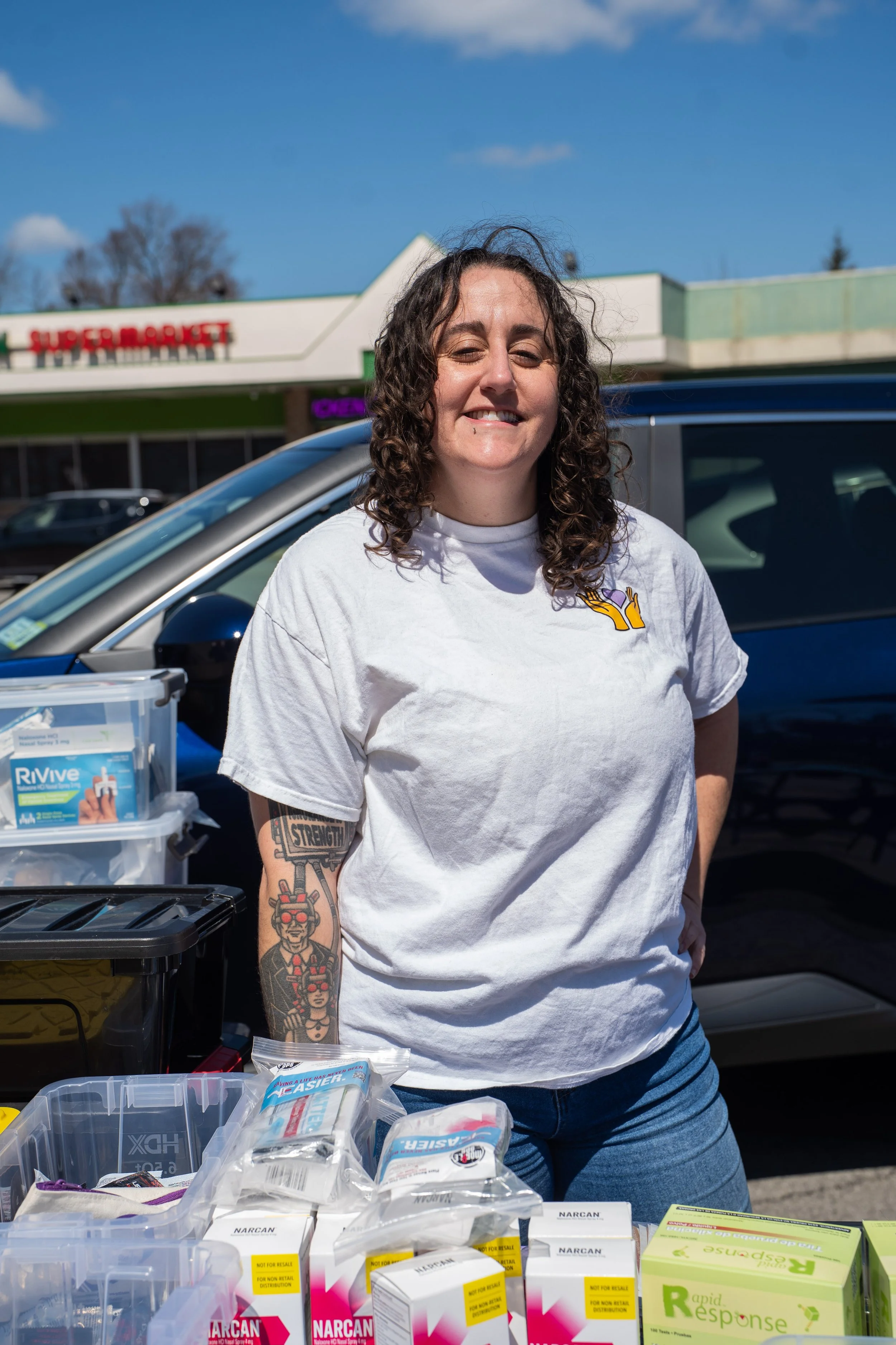 A smiling woman with curly brown hair, wearing a white T-shirt with a yellow hand logo, standing behind tables with medical supplies, outside in a parking lot with a supermarket in the background.