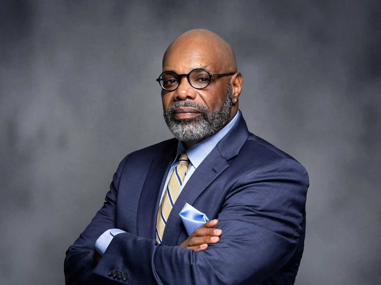 A confident African American man with a beard and glasses, wearing a navy suit, light blue shirt, yellow striped tie, and a pocket square, standing with arms crossed against a gray background.