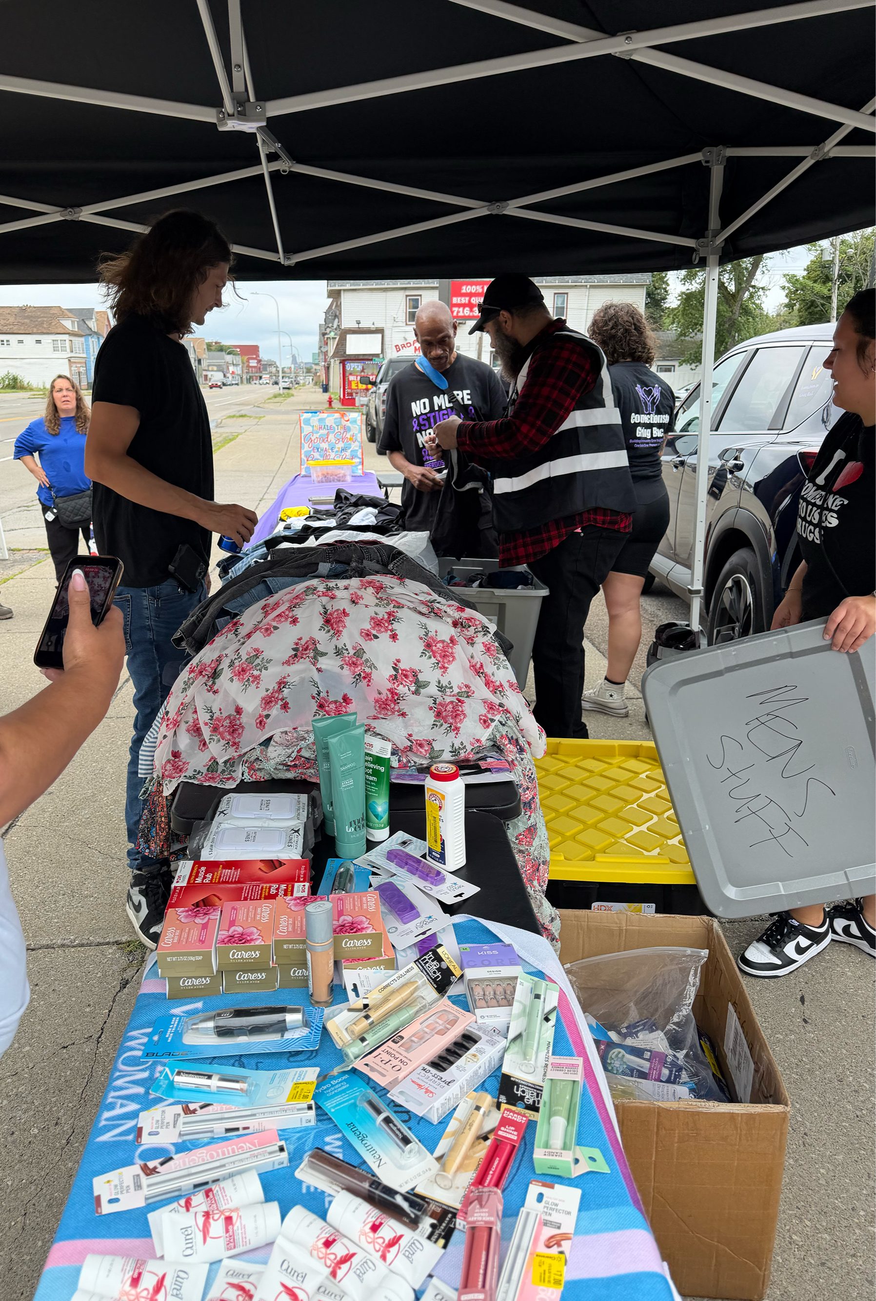 Outdoor table with products products for donation such as toiletries and cosmetics, under a black canopy, alongside people browsing and volunteering, with a street and buildings in the background.