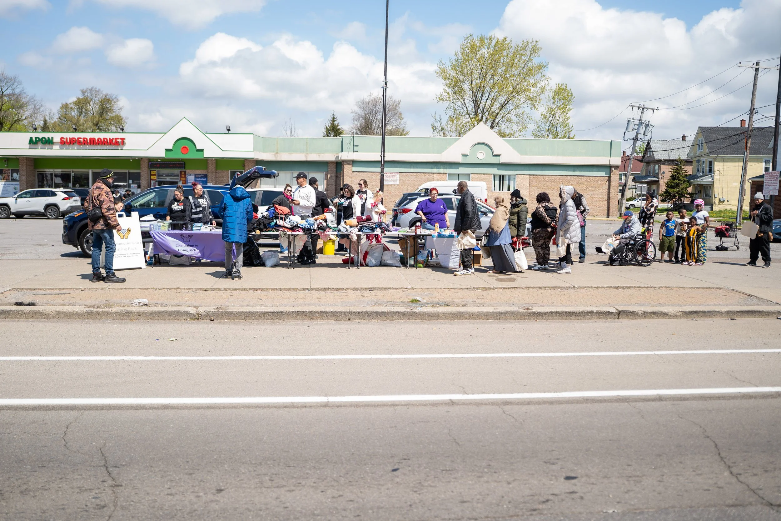 People gathered and shopping at an outdoor table of clothing and other items in front of a supermarket on a sunny day.