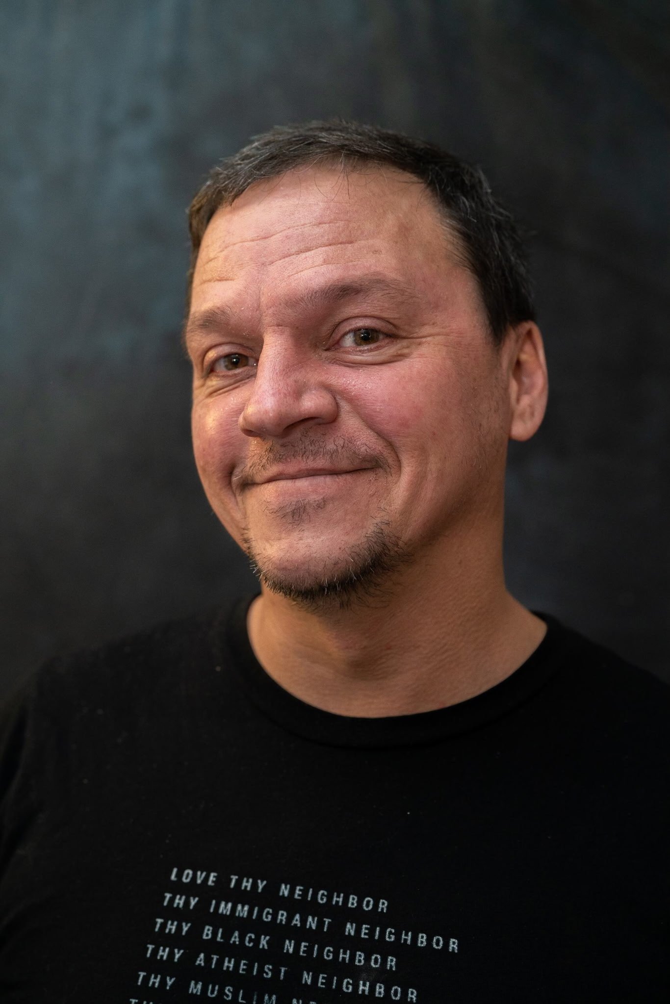 Close-up of a smiling man with short dark hair, light skin, and a small goatee, wearing a black shirt with text. The background is dark and the image is focused on his face and upper chest.