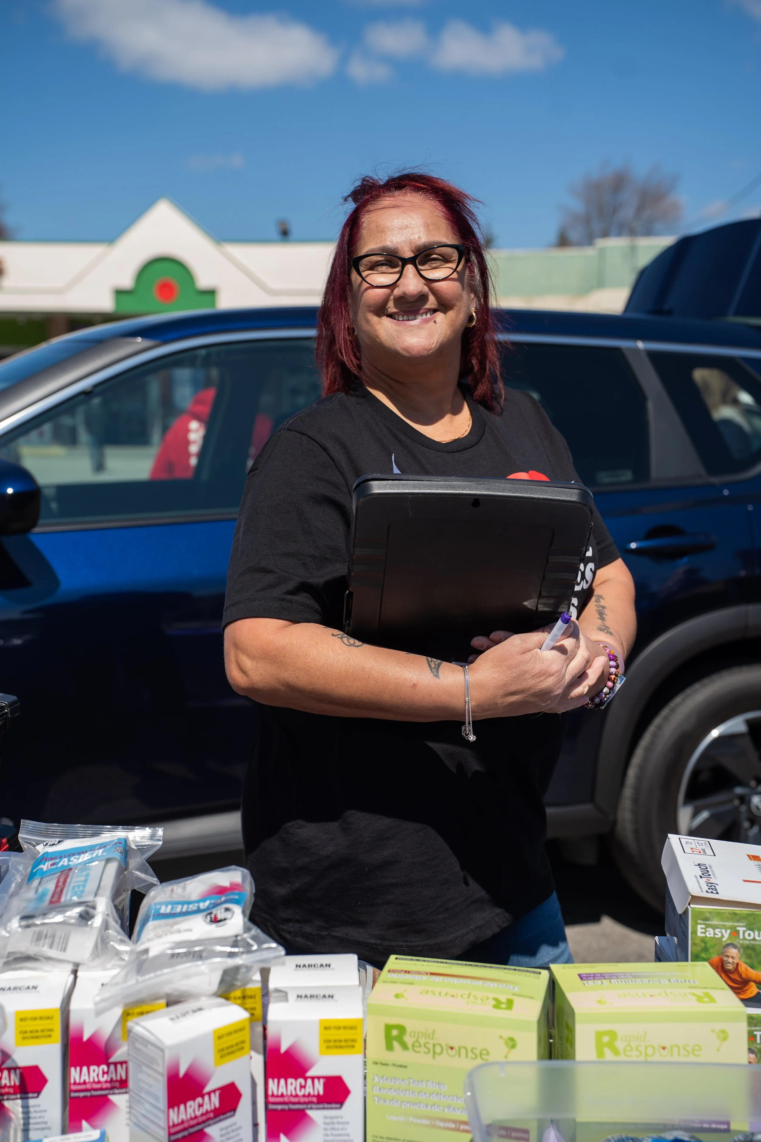 Smiling woman with red hair and glasses standing behind a table of medical supplies outdoors, with a blue sky and a building in the background.