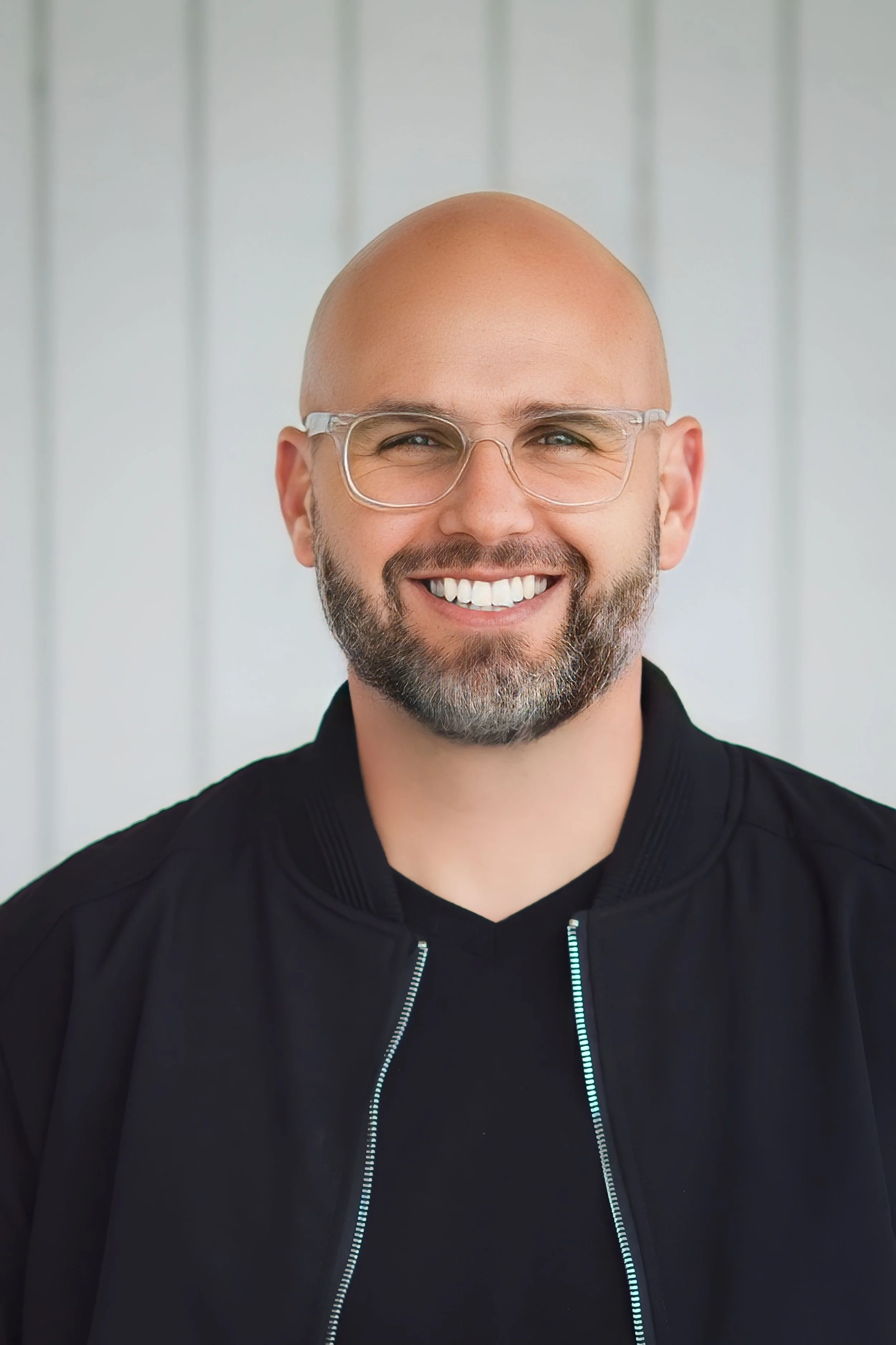 A smiling bald man with a beard wearing glasses and a black jacket, standing in front of a gray wall.
