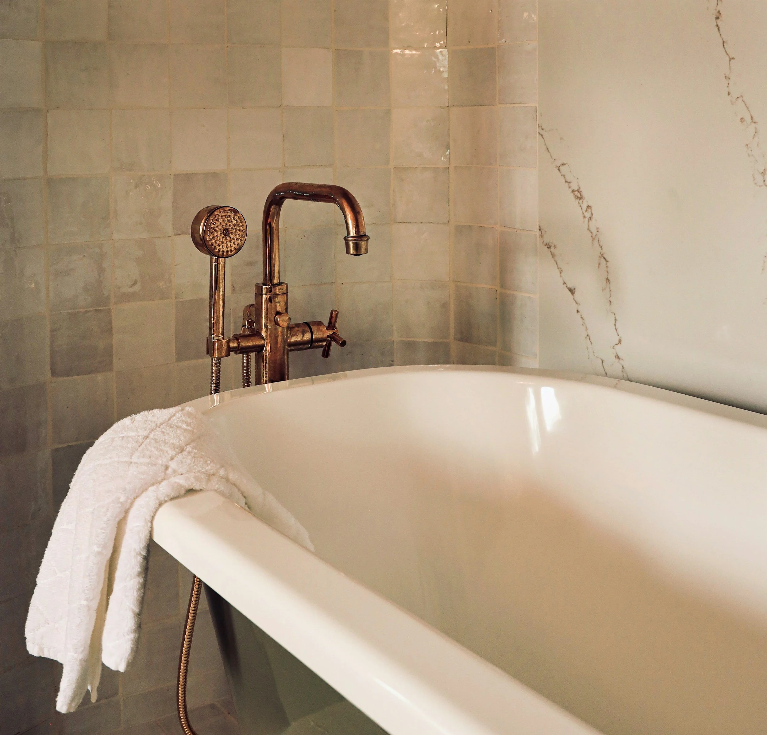 A white bathtub with a folded white towel on the edge, a vintage-style bronze faucet, a handheld shower head, a marble wall and corner, and teal-colored shelving in a bathroom.
