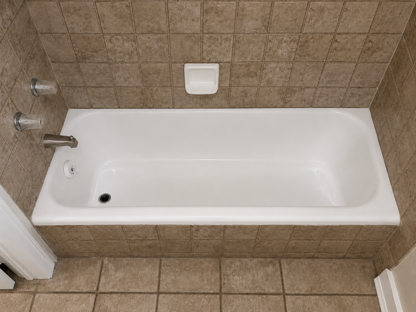 A white bathtub with a silver faucet, two clear glass knobs, a white soap dish mounted on tan tiled wall, and tan tiled floor.