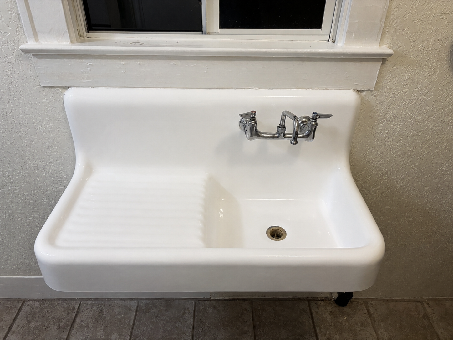 A white industrial utility sink with a chrome faucet, wall-mounted beneath a window in a room with textured off-white walls and gray tiled floor.