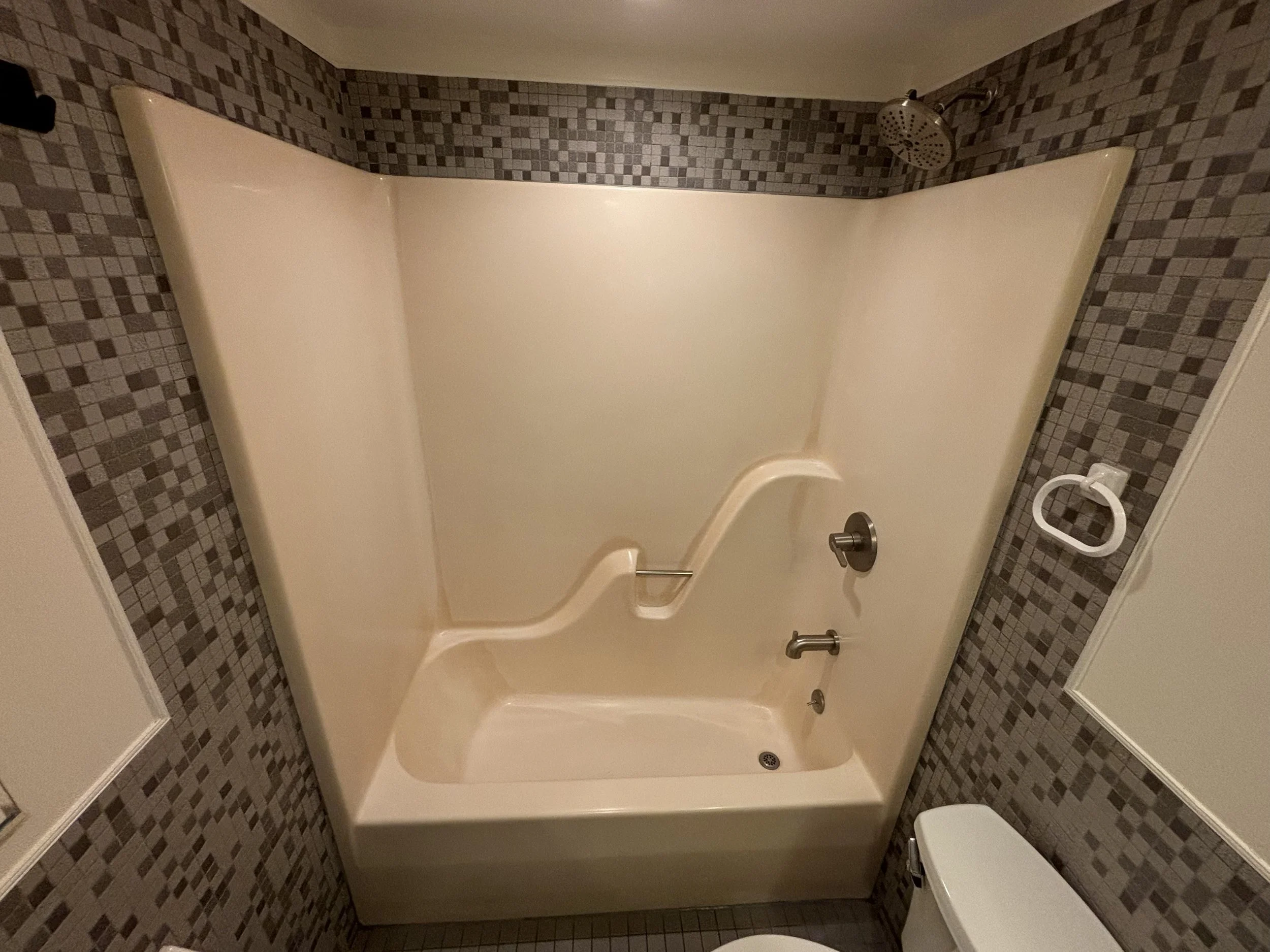Bathroom shower with beige tub, mosaic tile walls in shades of brown and gray, showerhead on the wall, and a white toilet at the bottom right corner.