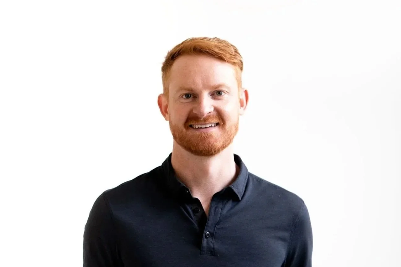 portrait of a smiling red-haired man with a beard, wearing a black collared shirt against a plain white background.