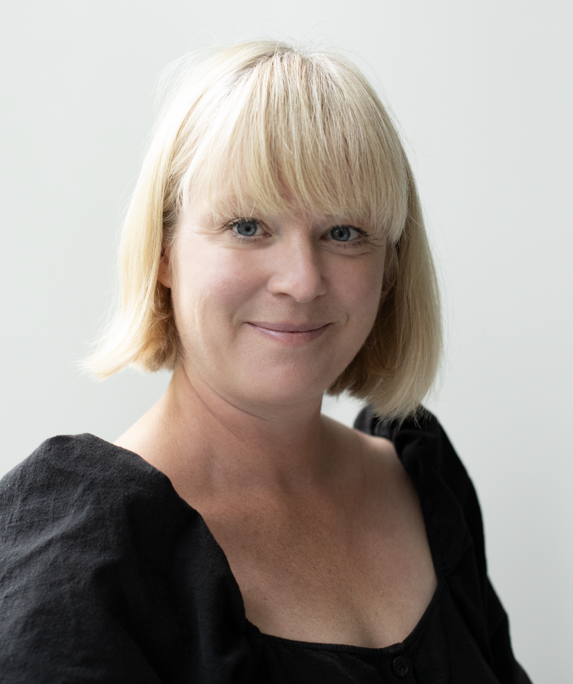 A woman with short blonde hair, blue eyes, and a gentle smile, wearing a black top with textured fabric, standing against a plain white background.
