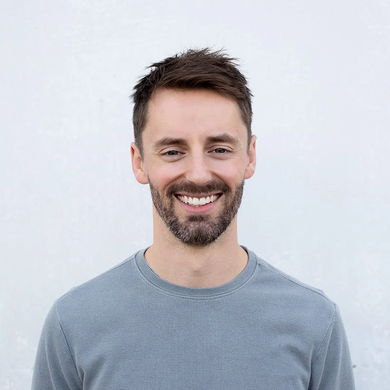 A man with brown hair, beard, and light skin smiling and wearing a gray shirt, standing against a plain light-colored background.