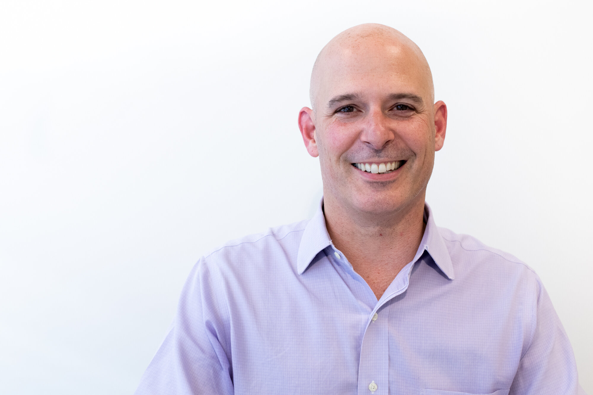 A smiling bald man in a light purple button-up shirt against a plain white background.