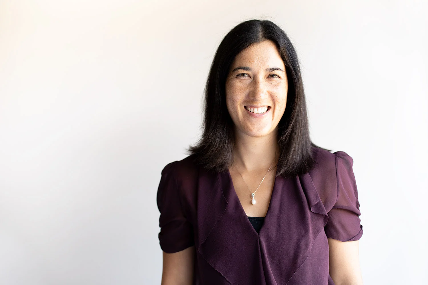 A woman with shoulder-length black hair smiling, wearing a purple blouse and a necklace, standing against a plain white background.