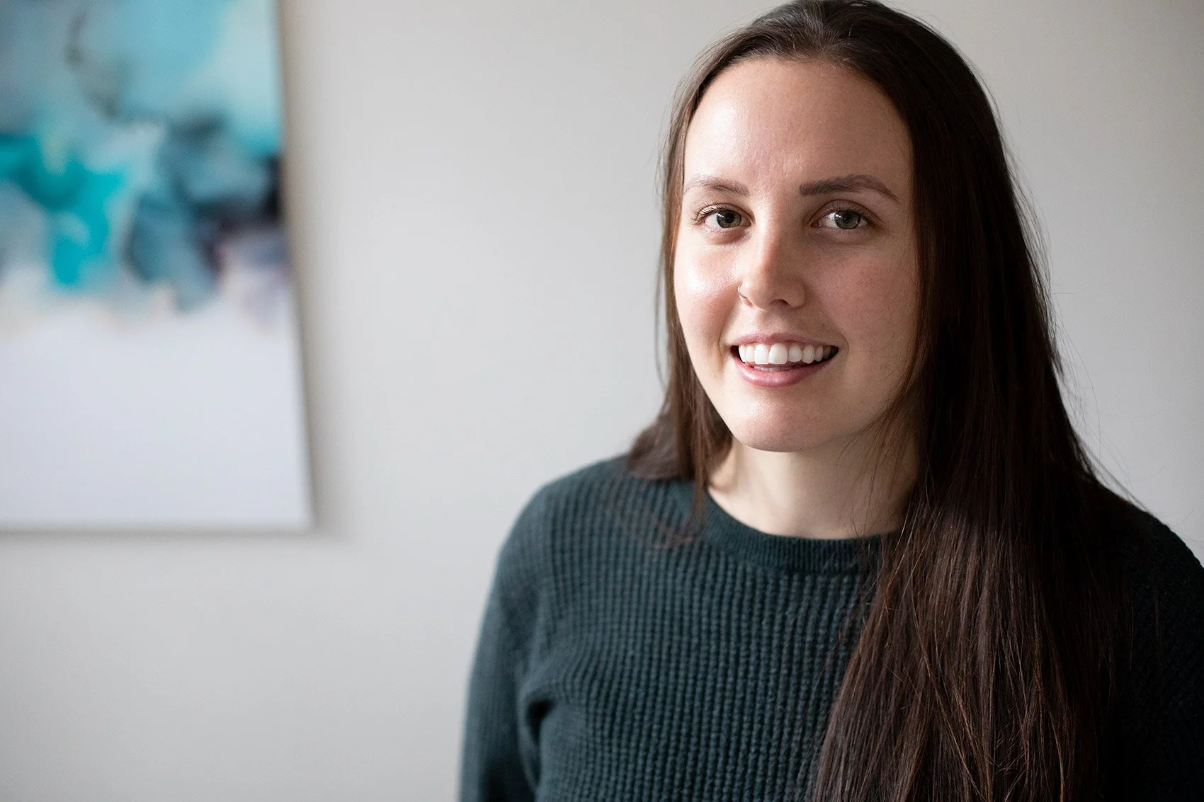 Close-up of a young woman with long, dark hair, smiling, wearing a dark green sweater, standing indoors with a white wall and an abstract artwork with blue and gray tones in the background.