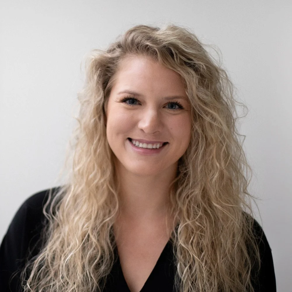 A woman with long, curly blonde hair smiling at the camera against a plain, light gray background.