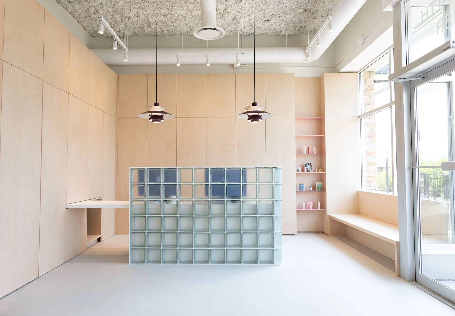 Bright modern interior with beige wooden wall panels, large windows, and ceiling spotlights. There are two pendant lamps hanging from the ceiling, a glass block partition, pink shelves with books, and a built-in seating area beneath the windows.
