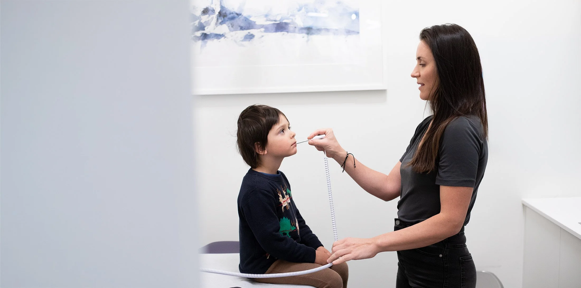 A woman with long dark hair wearing a black shirt performs a medical examination on a young boy with dark hair, wearing a sweater with Christmas trees, in a clinical setting with white walls and a watercolor painting in the background.