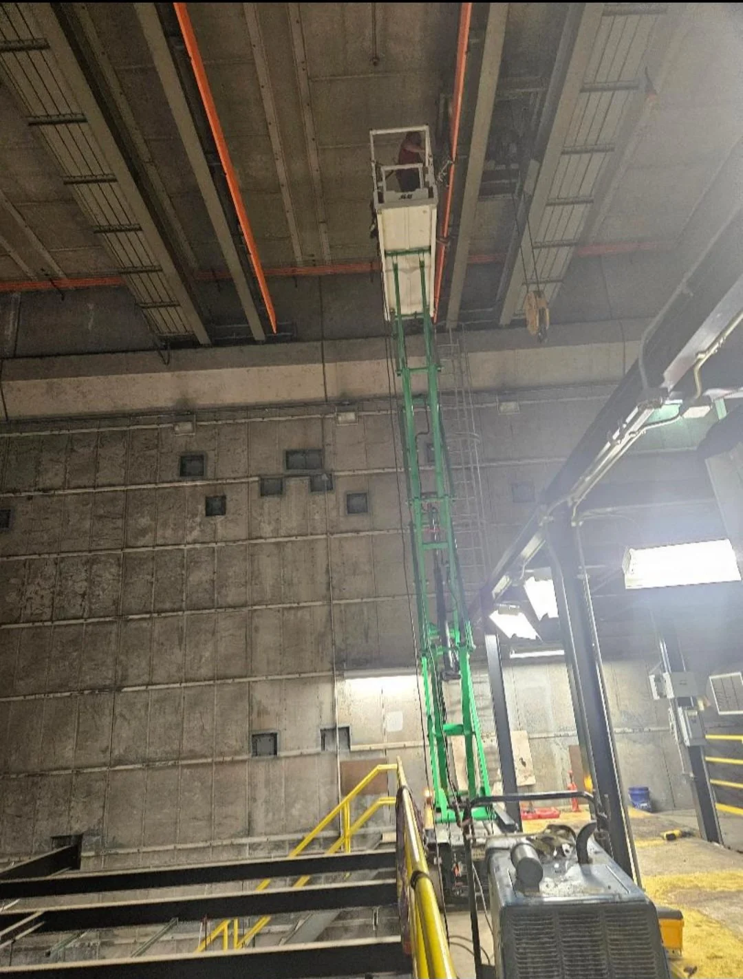 An industrial lift or cherry picker extending towards the ceiling of a large concrete warehouse or parking garage. The ladder is green, and the platform at the top appears to be used for maintenance purposes.