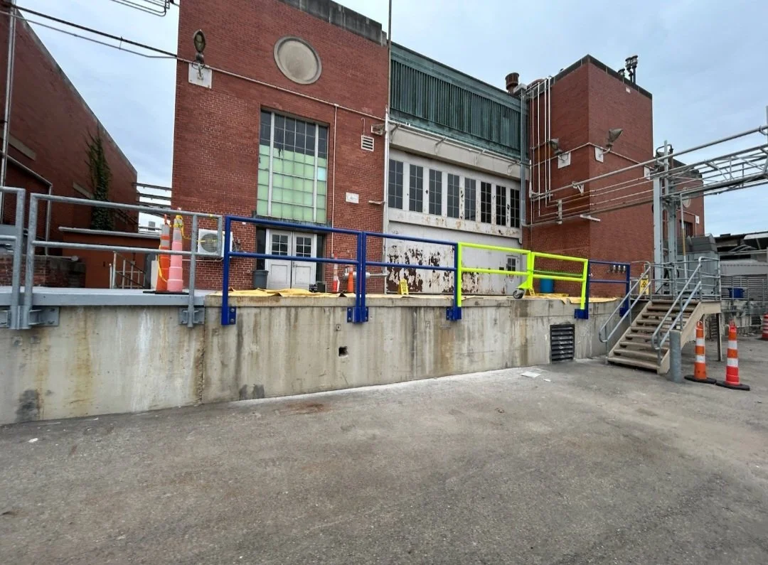 Exterior view of an industrial building with safety railings, orange cones, stairs, and utility pipes.