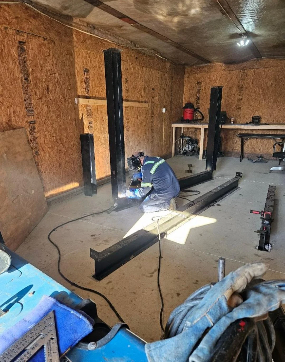 A worker welds metal beams in a construction site indoor space with plywood walls, a workbench, and construction tools scattered around.