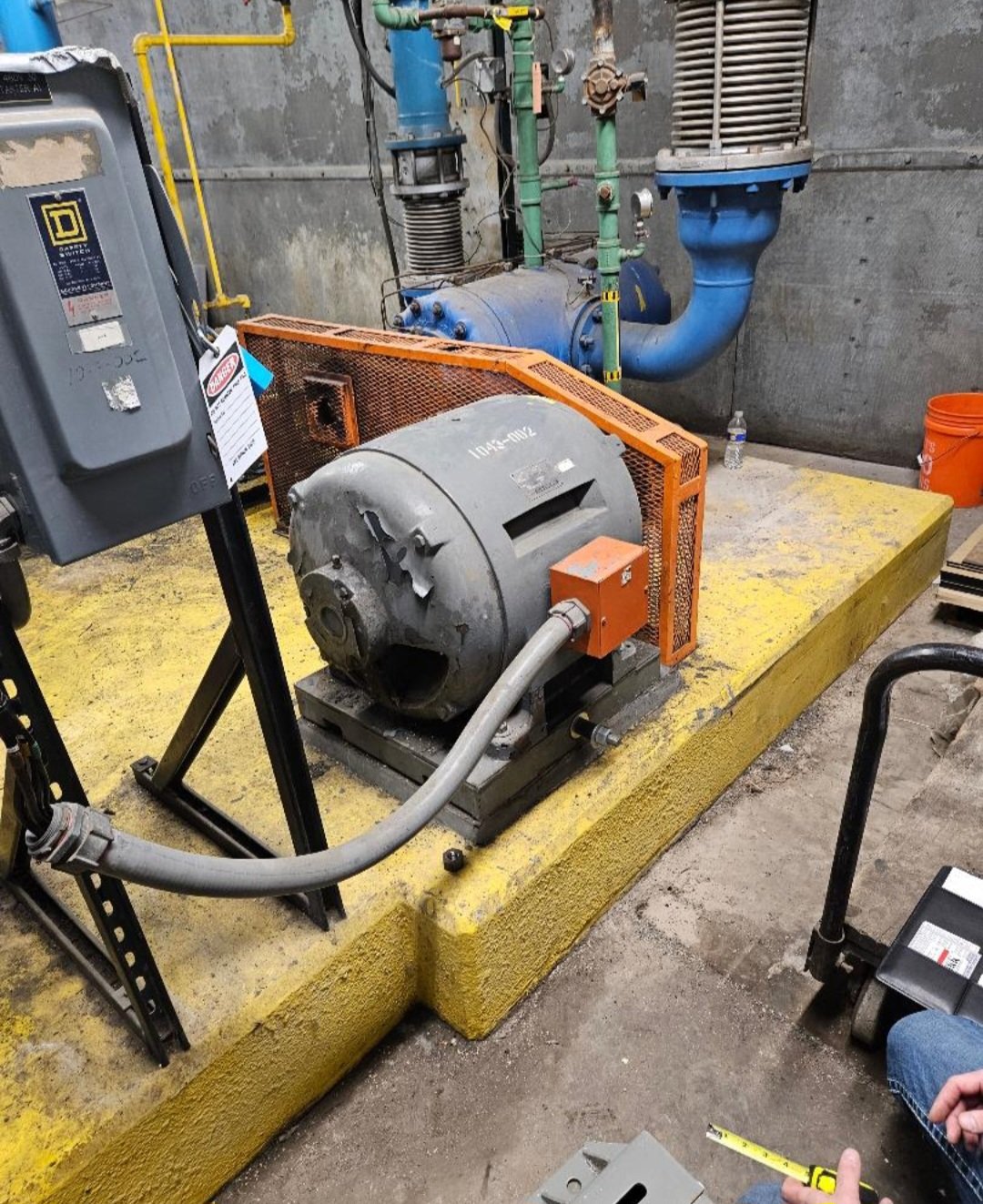 Industrial mechanical equipment including a large motor with a hose attached, an orange safety guard, electrical box, and various pipes and valves in a utility room.