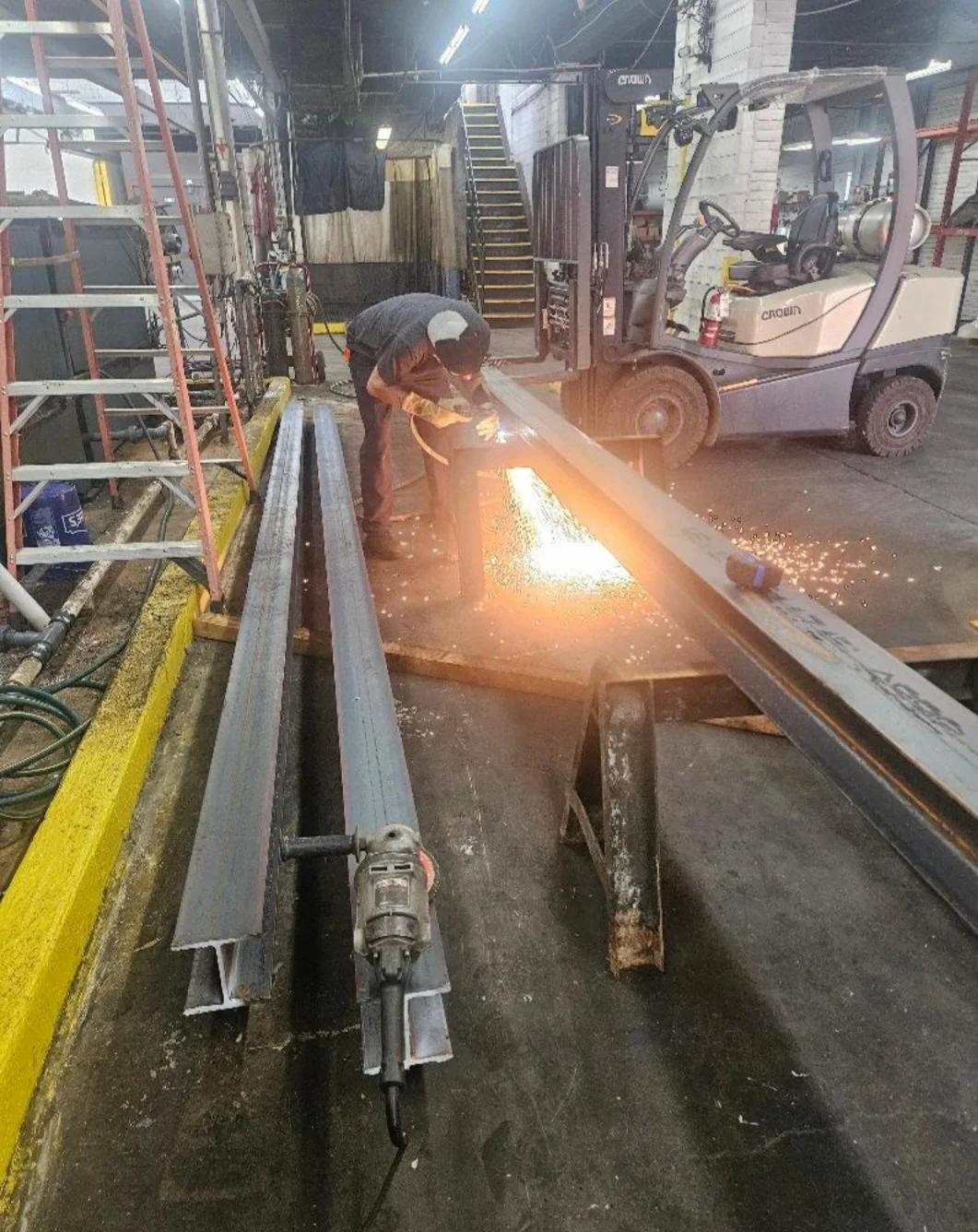 A worker welding metal beams in an industrial workshop, with sparks flying, a forklift nearby, and various tools and equipment around.