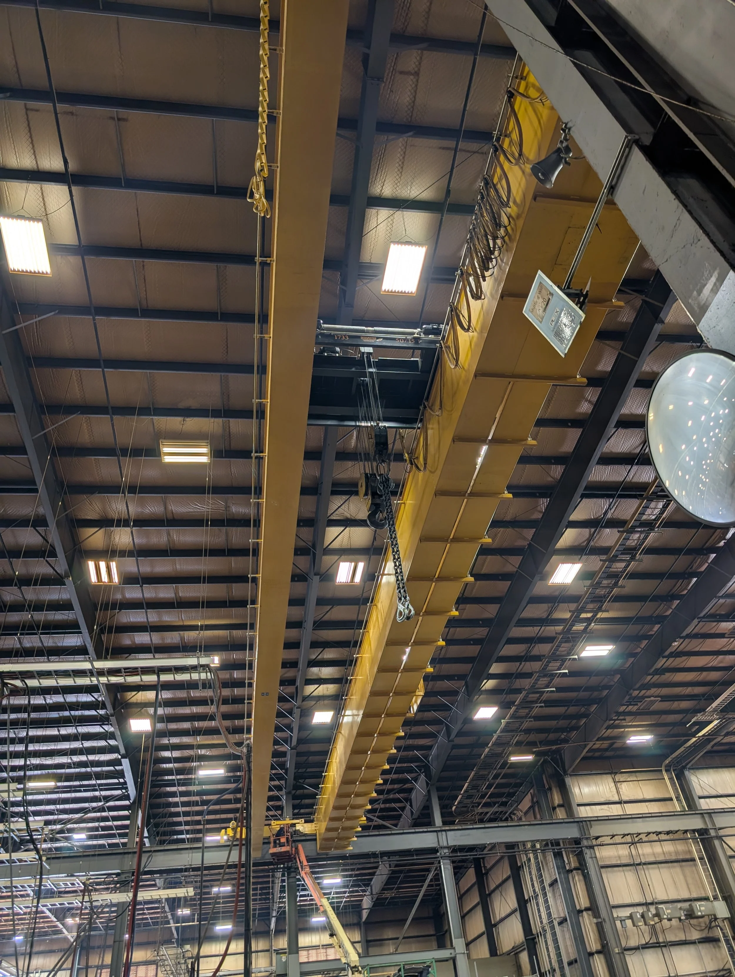 Inside of an industrial warehouse with a large overhead crane system, featuring a yellow beam and hoist, with various electrical and mechanical equipment and lighting overhead.