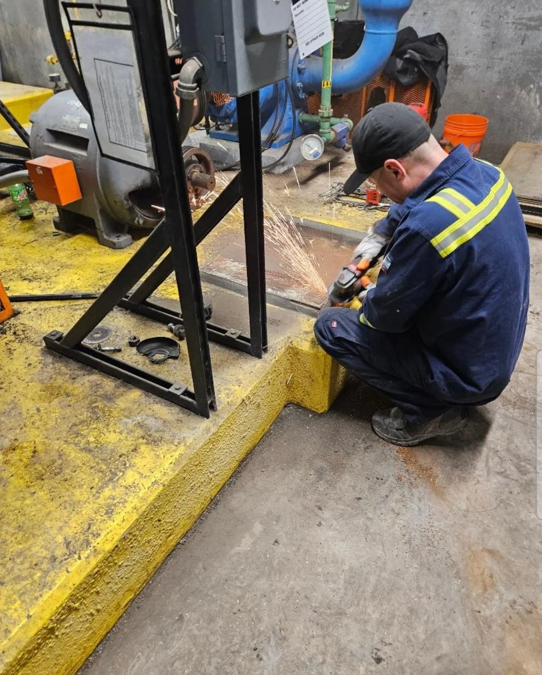 A worker in safety gear using a grinder to cut or grind metal, with sparks flying, in an industrial workshop setting.