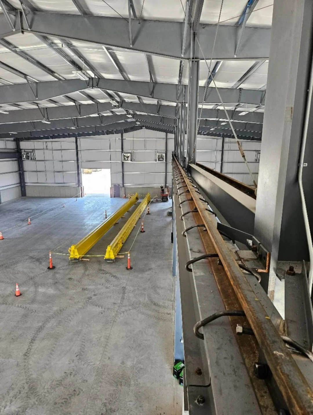 Interior of a large industrial warehouse with a high ceiling and open space, featuring yellow overhead cranes, orange safety cones, and construction equipment.