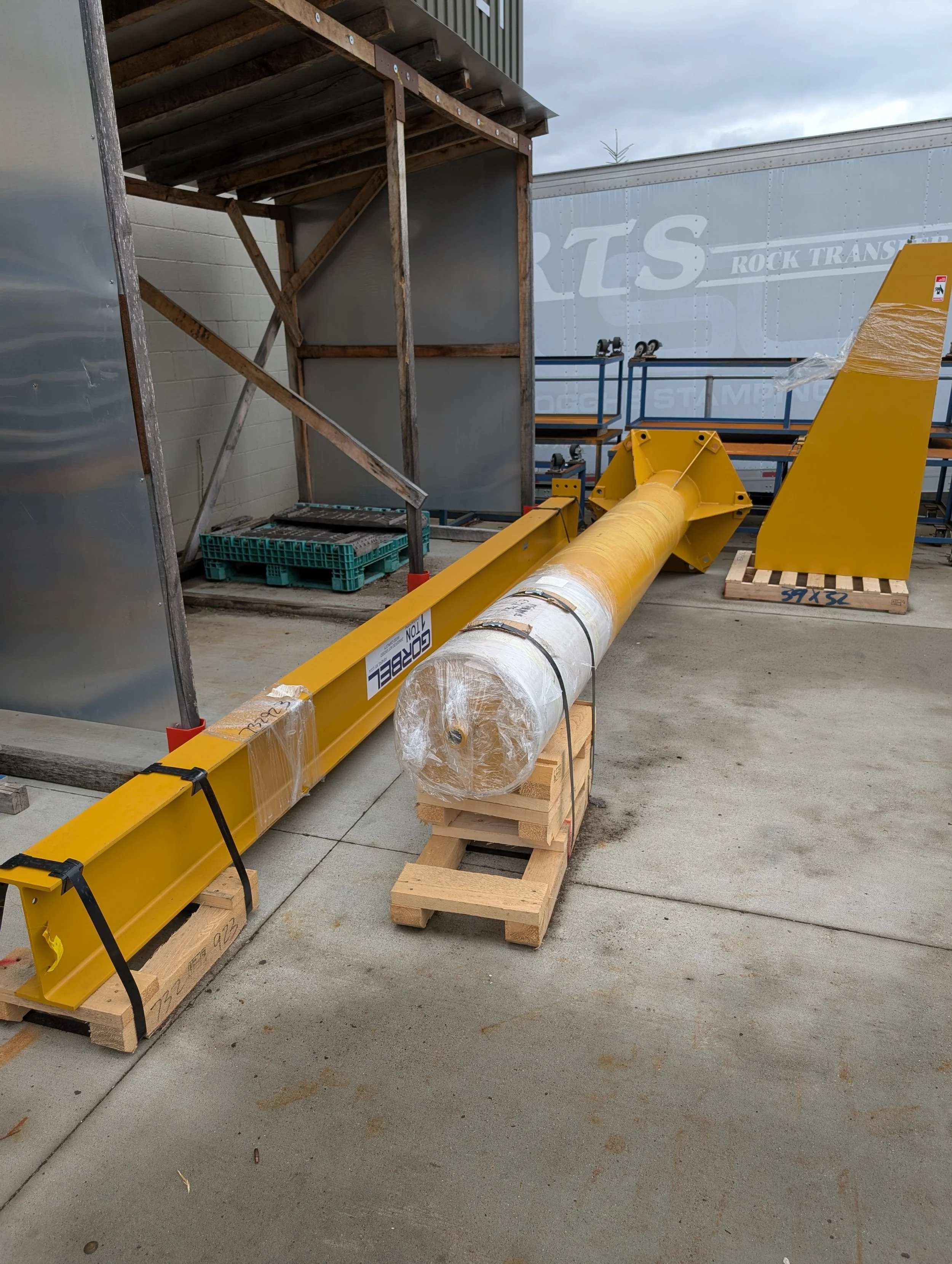 Construction equipment including a yellow safety barrier and a large pipe placed on wooden pallets outside a building, with a large truck in the background and a cloudy sky.