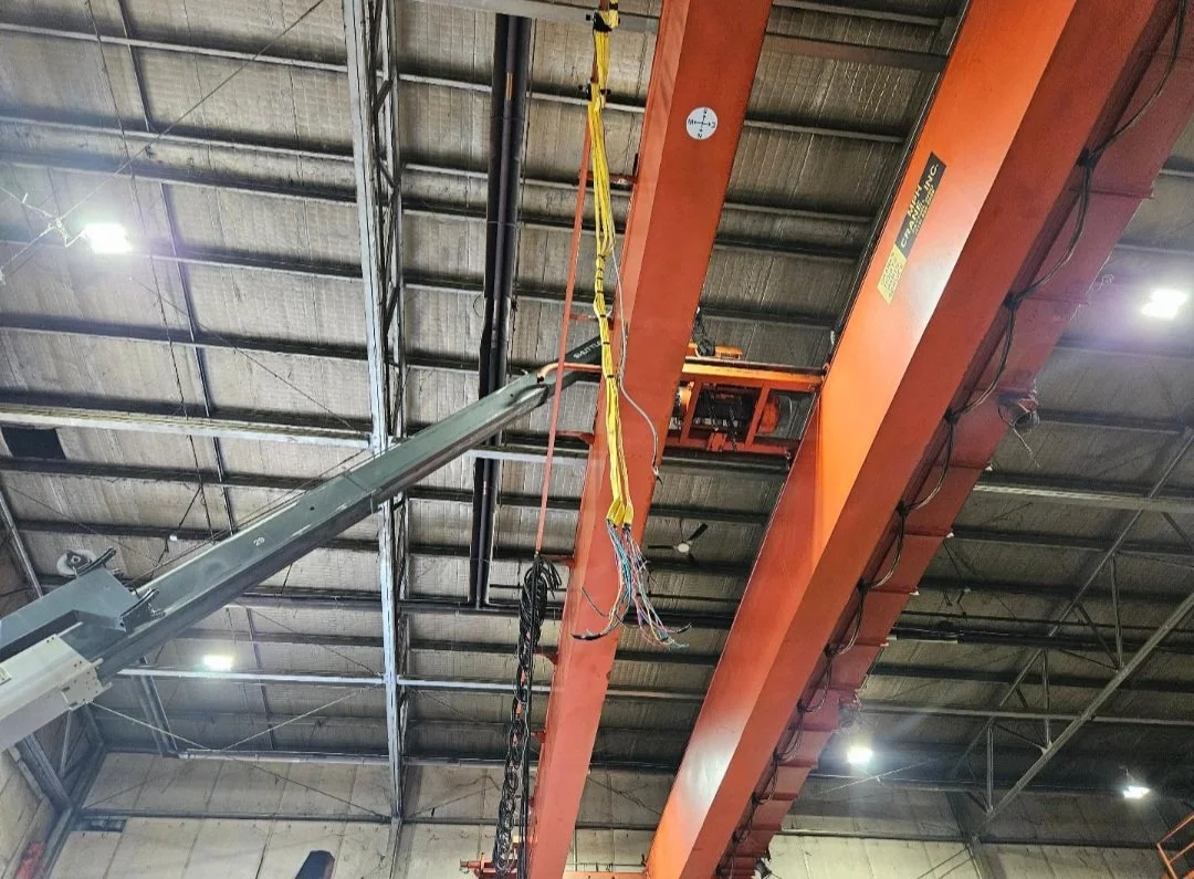 Inside a factory or warehouse with a large overhead crane and electrical wires hanging from the ceiling.