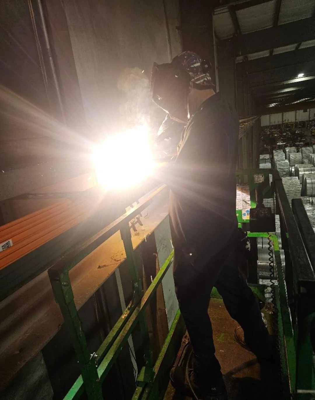 A person welding inside a factory or warehouse, wearing protective gear including a helmet and apron, with bright sparks from the welding process illuminating the scene.