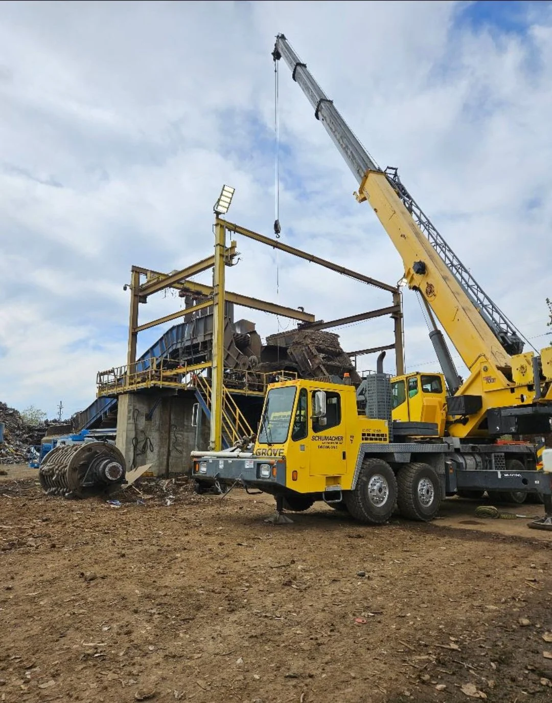 Construction site with yellow crane lifting metal framework, industrial machinery, and a dirt ground.