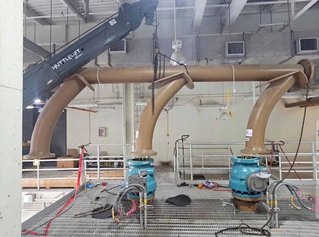 Industrial setting with large brown pipes connected to blue machinery, surrounded by metal grating, tools, and equipment.