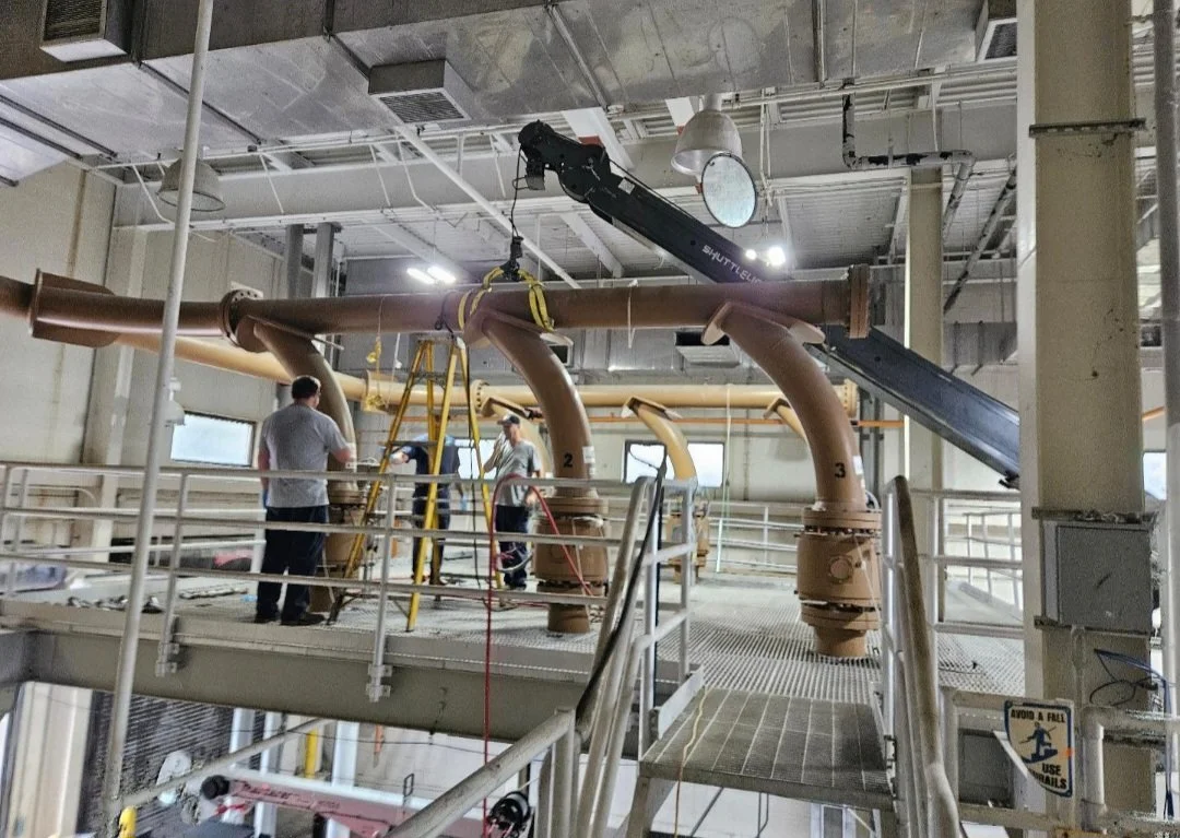 Industrial interior with large brown pipes and elevated walkways, workers inspect equipment, and a crane aids in maintenance or installation.