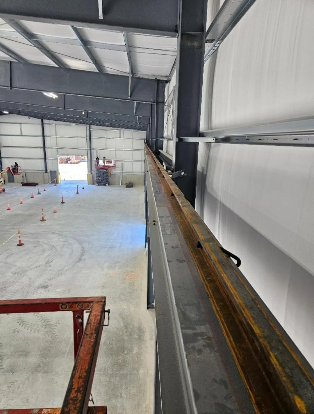 Inside of a large industrial warehouse with a steel structure. Construction equipment and safety cones are visible on the floor, and a lift platform is present in the foreground.