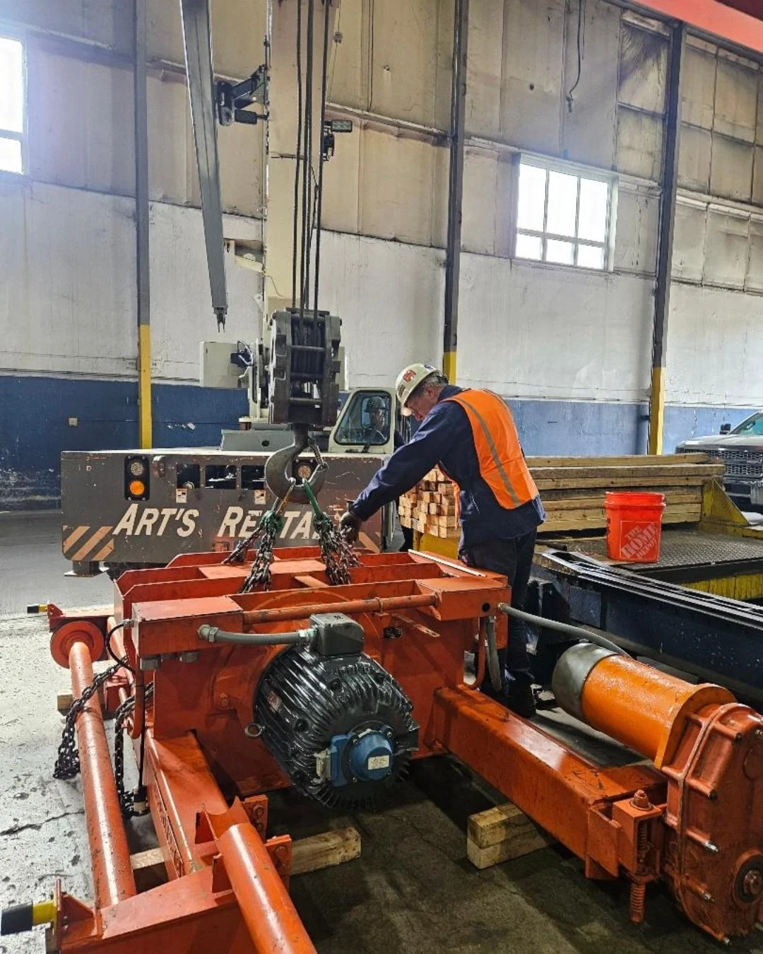 A worker wearing a safety helmet and orange safety vest is working with a large orange industrial machine inside a warehouse. The worker appears to be adjusting or inspecting the equipment, which is mounted on a wooden pallet, with stacks of lumber a