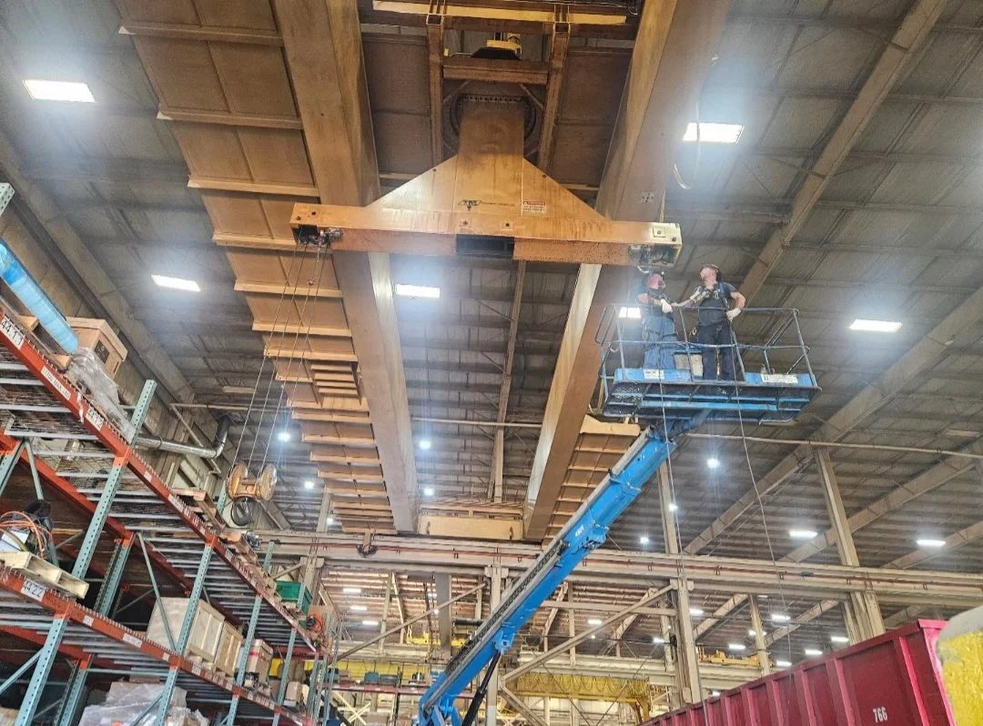 Two workers on a blue lift working on a large overhead industrial crane inside a warehouse or manufacturing facility.