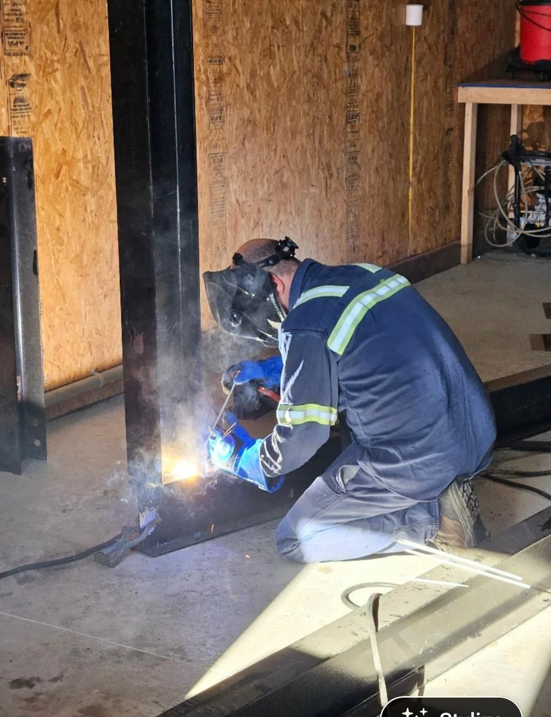 A person wearing a welding mask and protective gloves is welding a metal post inside a wooden construction area.