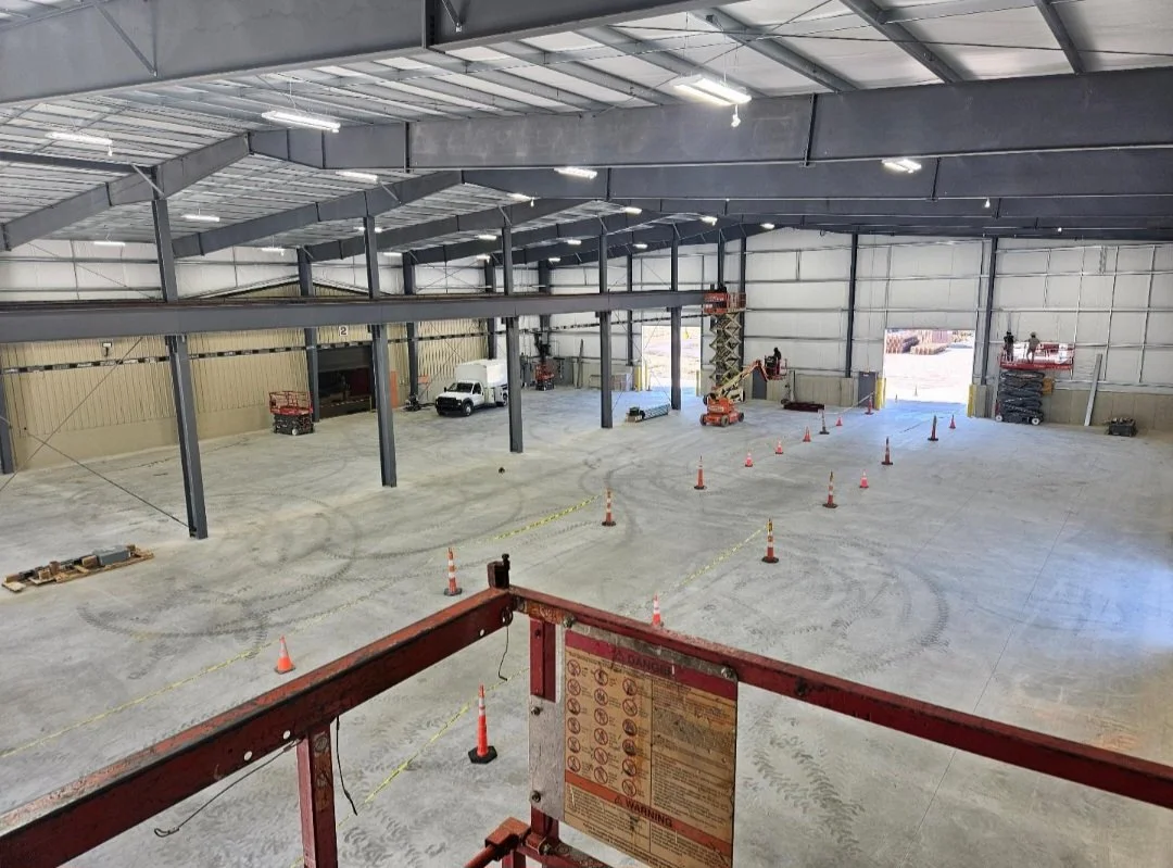 Inside a large industrial warehouse under construction, with construction equipment, orange safety cones, and a white truck parked inside.