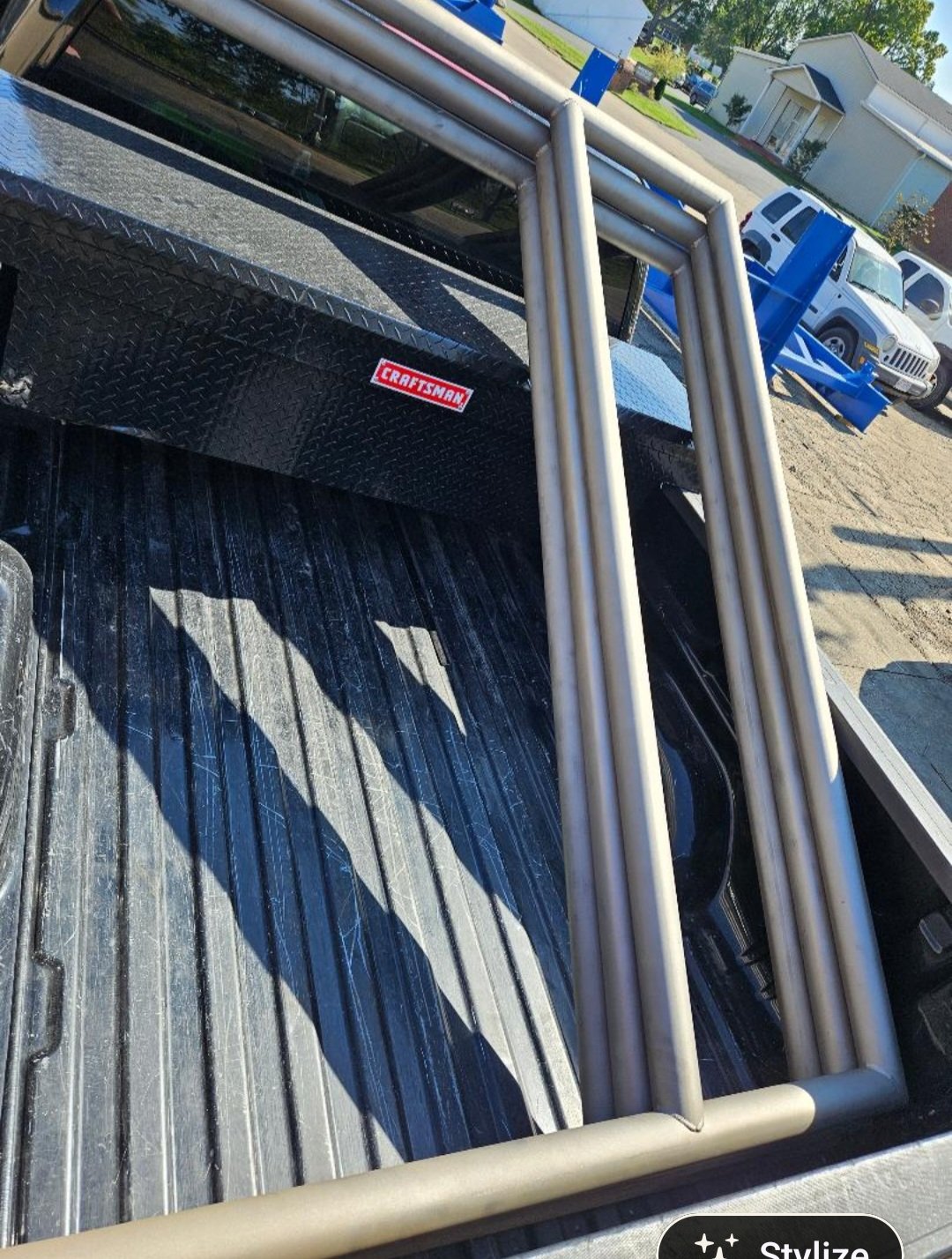 The back of a pickup truck with a metal frame and a black toolbox with a Craftsman logo, parked on a street with houses and vehicles in the background.